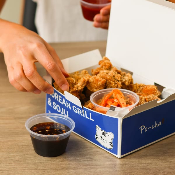 Hand reaching into a Po-cha box of Korean fried chicken with kimchi and dipping sauce.