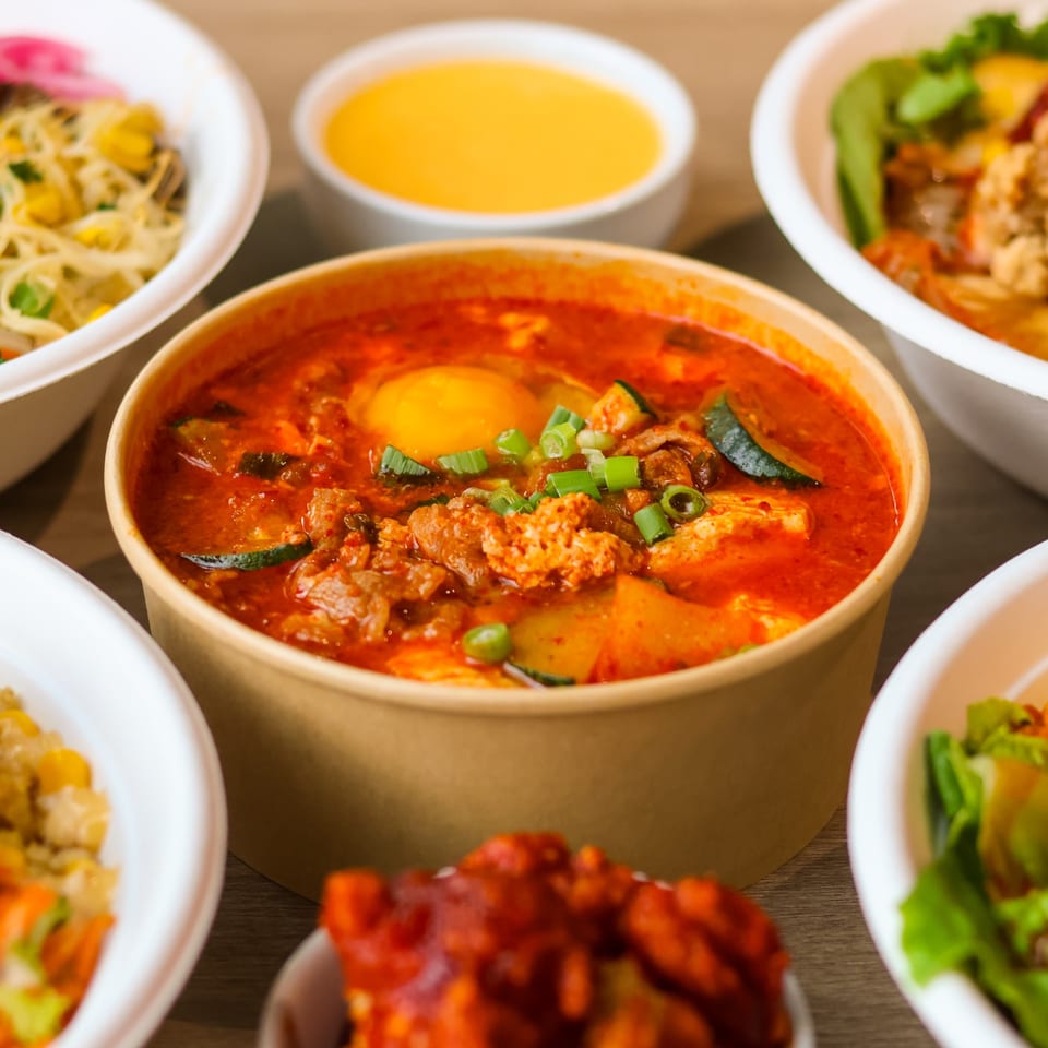 Spicy red soup with an egg and green onions, surrounded by various takeout dishes.