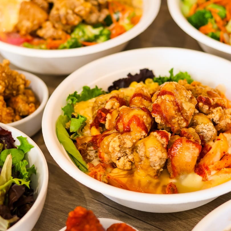 Central bowl of fried chicken with orange and red sauces, lettuce, and corn, surrounded by other dishes.