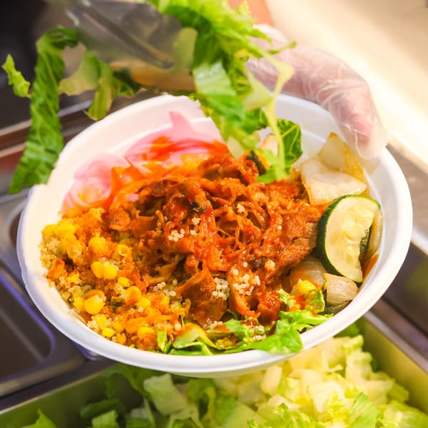 Gloved hand adds fresh lettuce to a vibrant bowl of pulled pork, quinoa, corn, and vegetables.