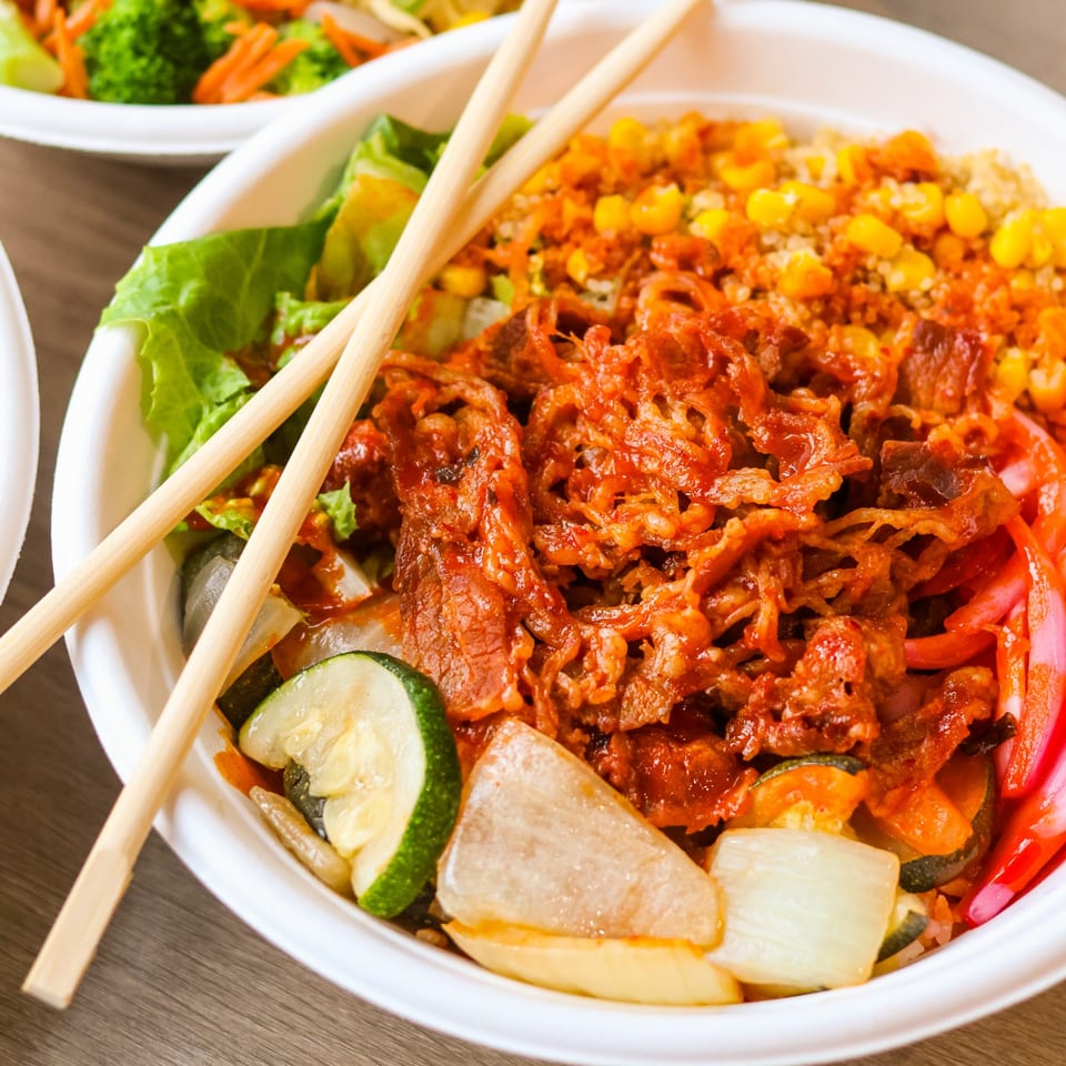Vibrant bowl of Asian-inspired food with red sauced meat, corn, vegetables, and chopsticks.
