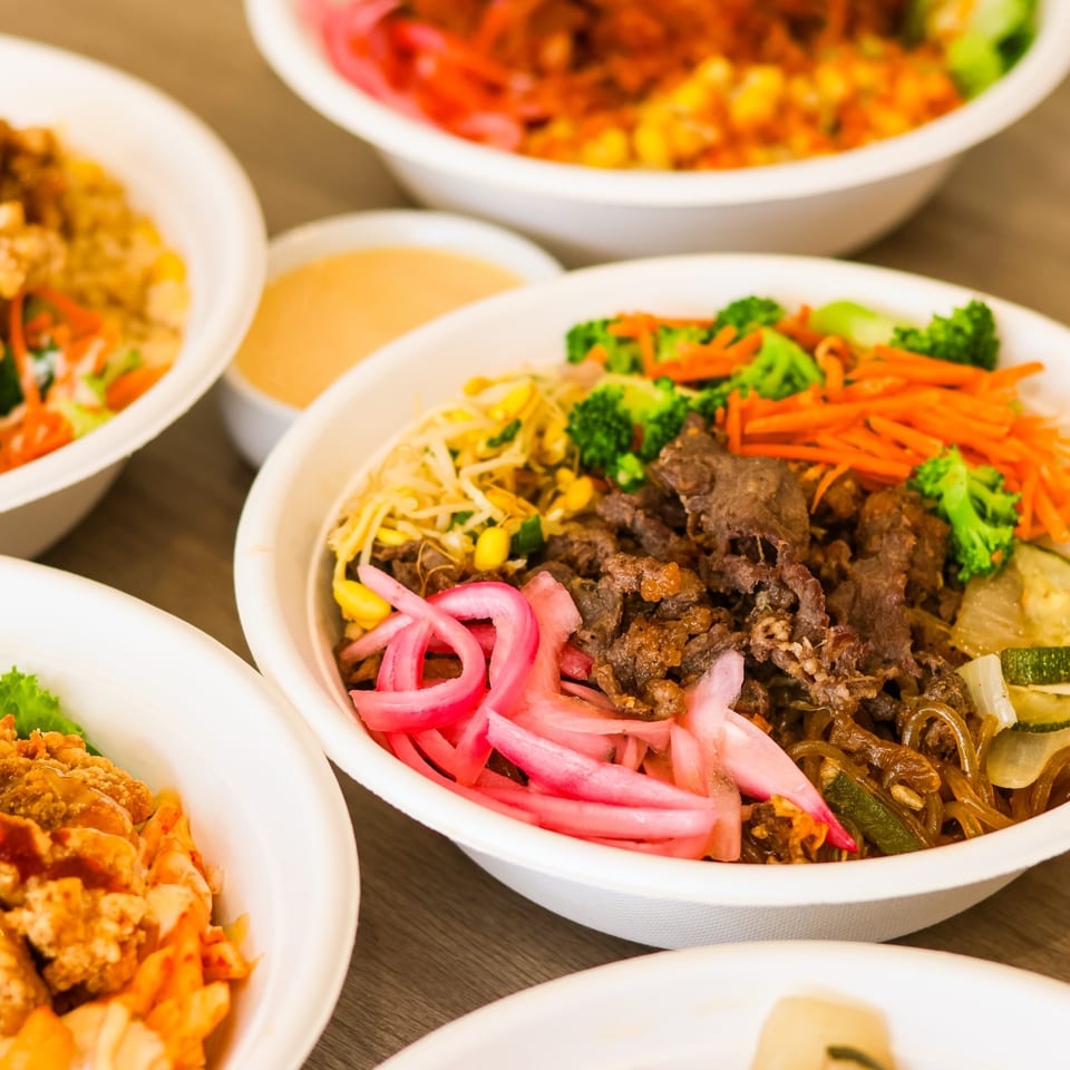 Several colorful Korean-style bowls with beef, pickled onions, vegetables, and noodles on a wooden table.