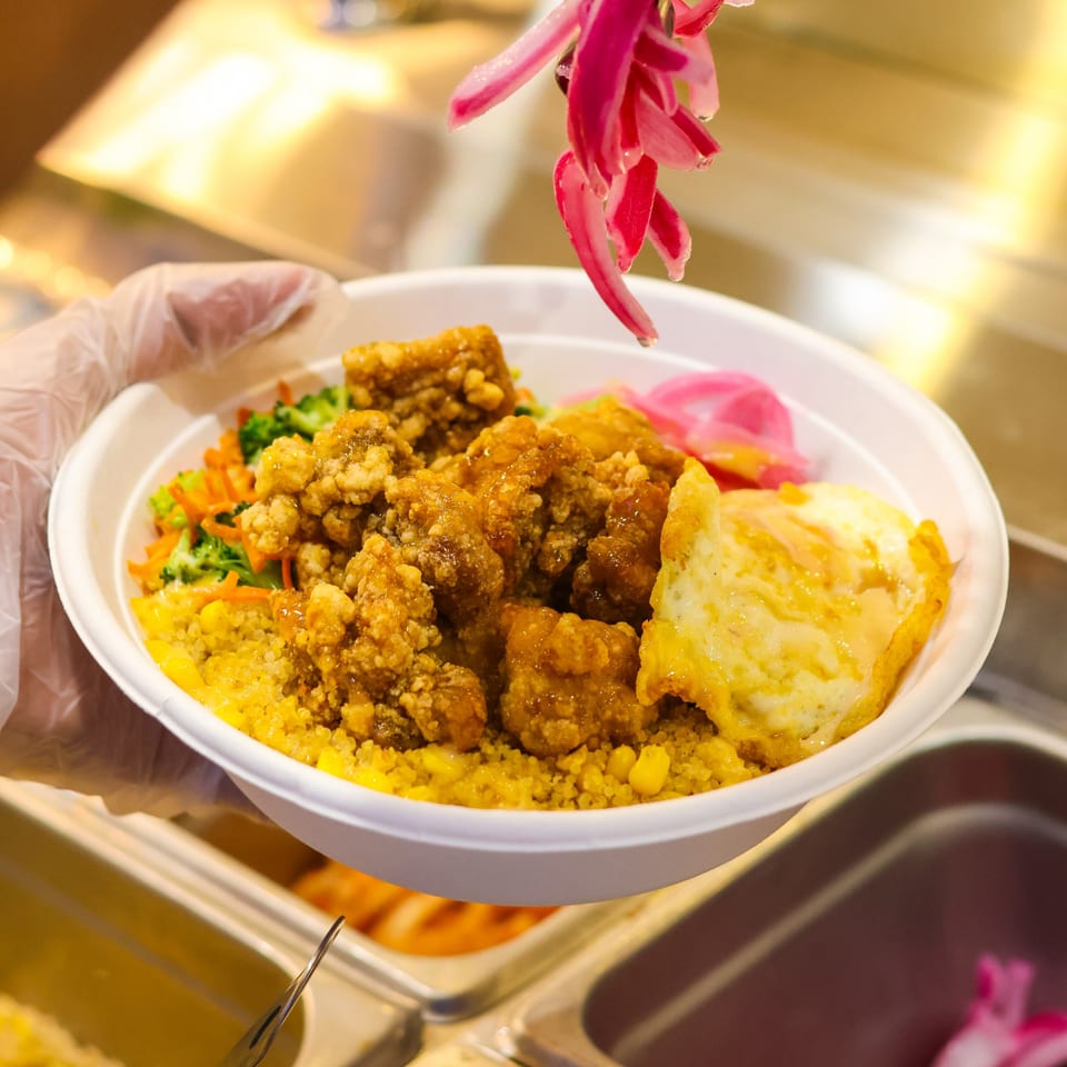 A gloved hand presents a bowl of fried chicken, yellow rice, fried egg, and pickled onions.