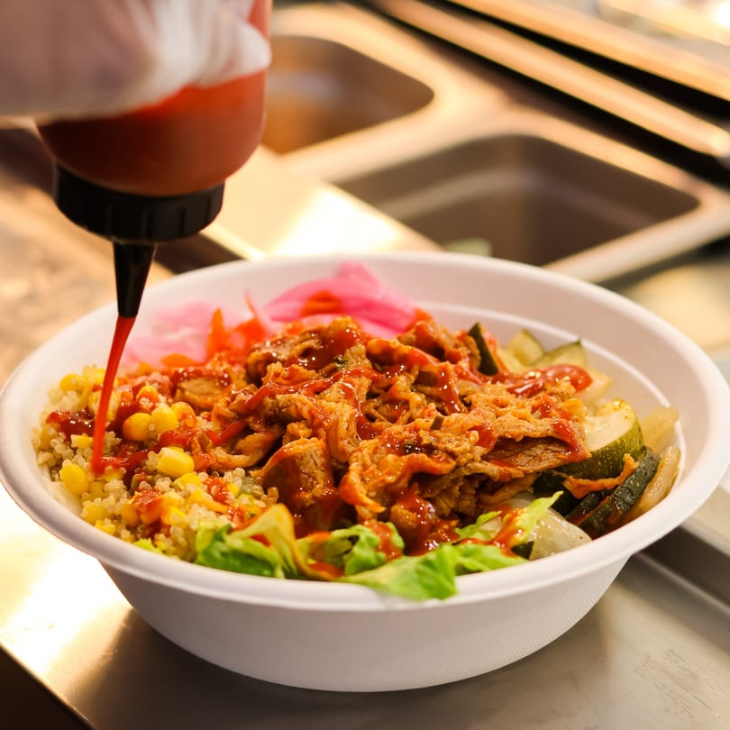 Red sauce drizzled onto a vibrant bowl of quinoa, meat, corn, and pickled vegetables.