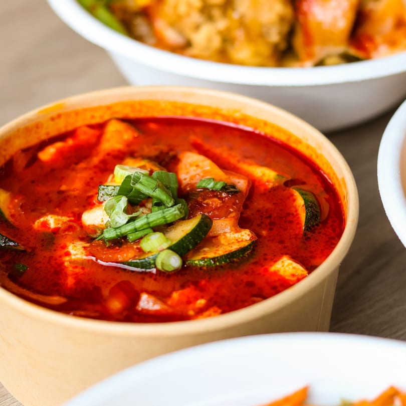 Red soup with zucchini, tofu, and green onions in a takeout container.