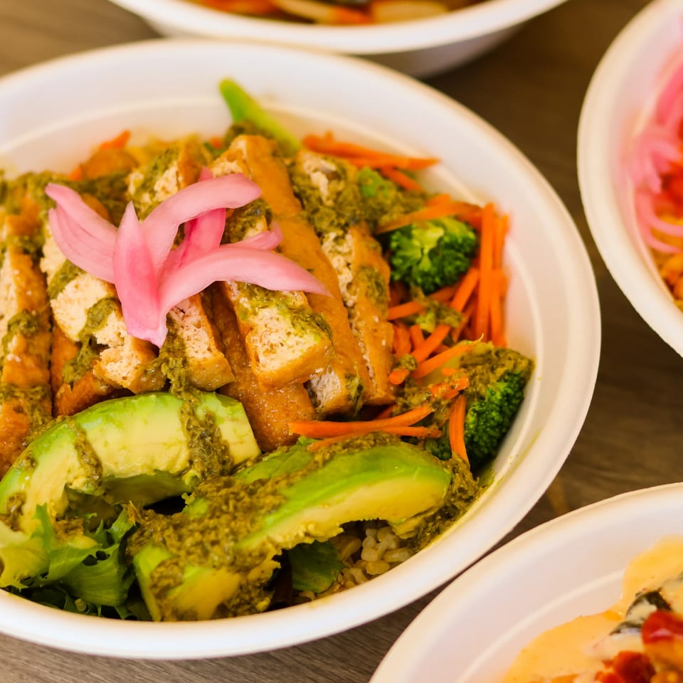 A vibrant bowl of tofu, avocado, broccoli, carrots, and pickled onions with green sauce.
