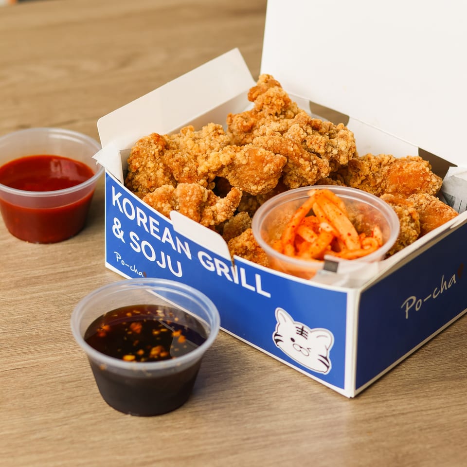 Crispy Korean fried chicken in a Po-cha box with kimchi and two dipping sauces on wood.