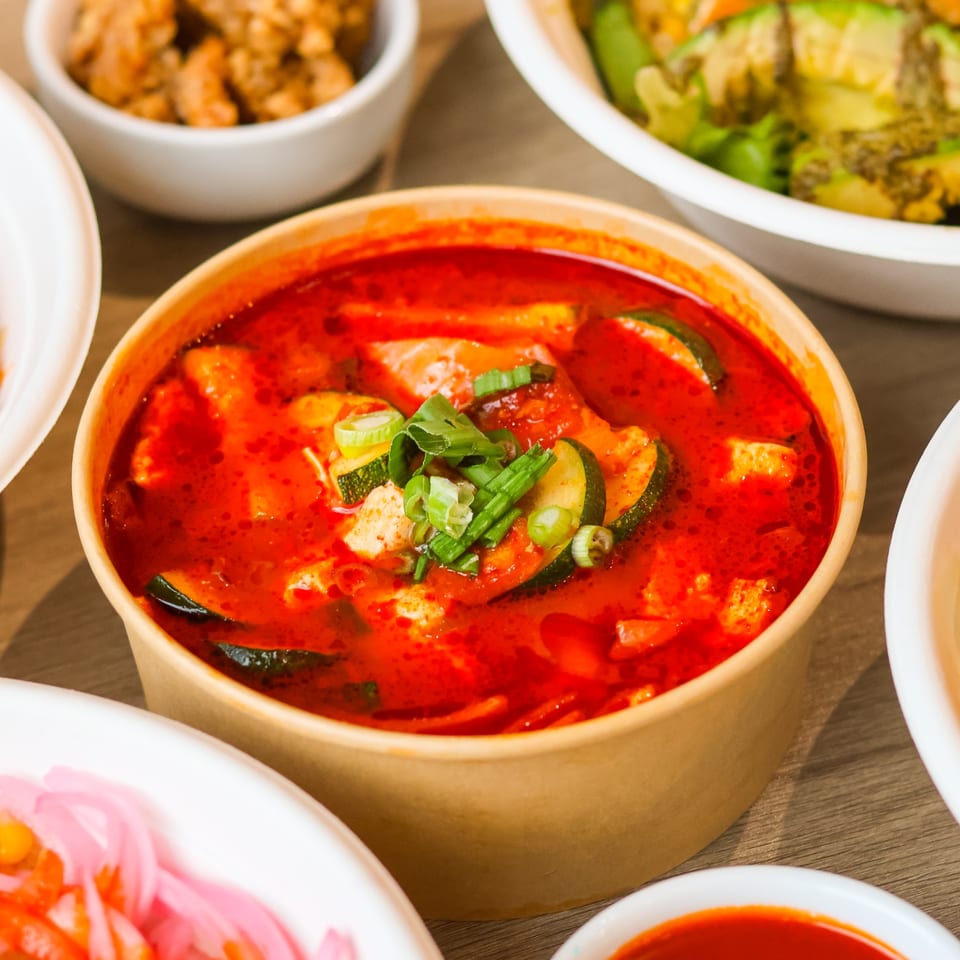 A vibrant red soup with zucchini, tofu, and green onions in a brown bowl, surrounded by other dishes.