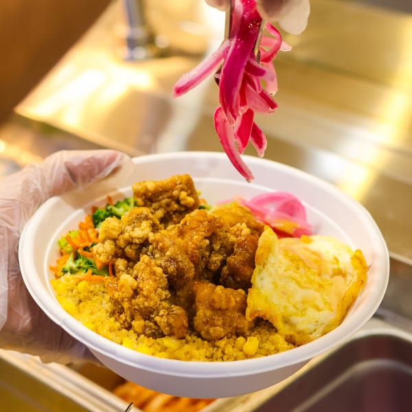 Gloved hand adds pickled red onions to a fried chicken rice bowl with vegetables and egg.