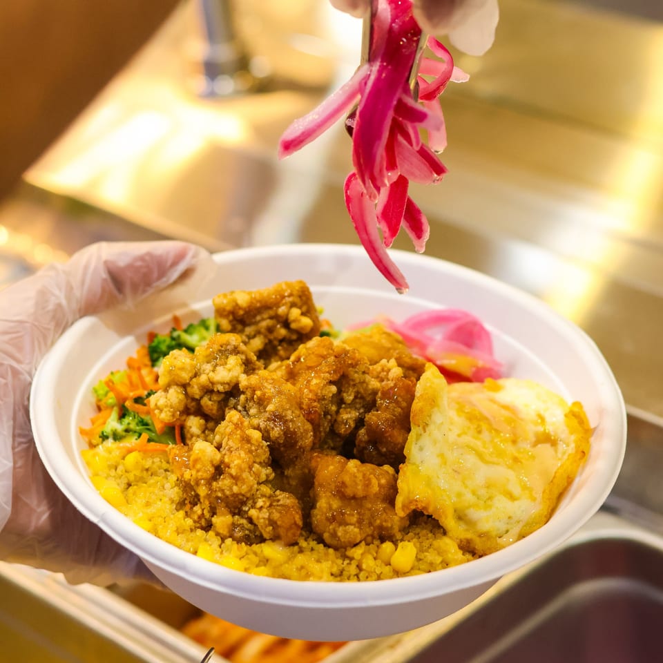Gloved hand adds pickled red onions to a fried chicken rice bowl with vegetables and egg.