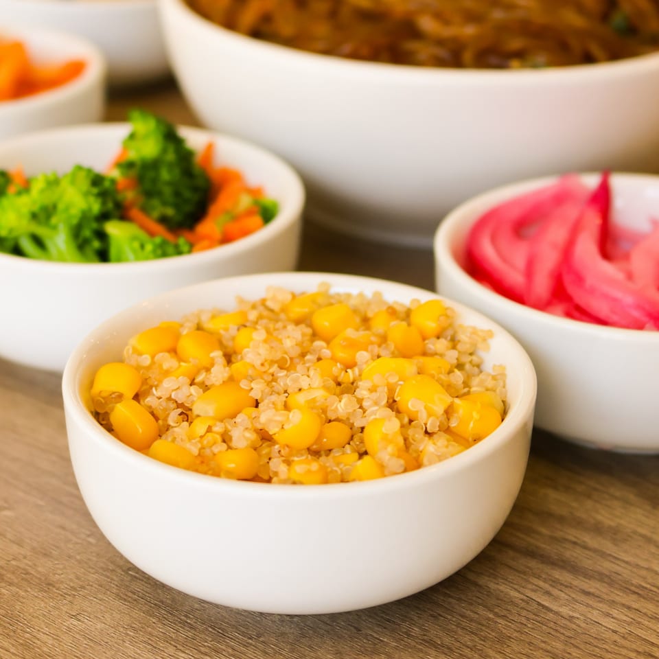 White bowl of quinoa and corn with other bowls of vegetables on a wooden table.